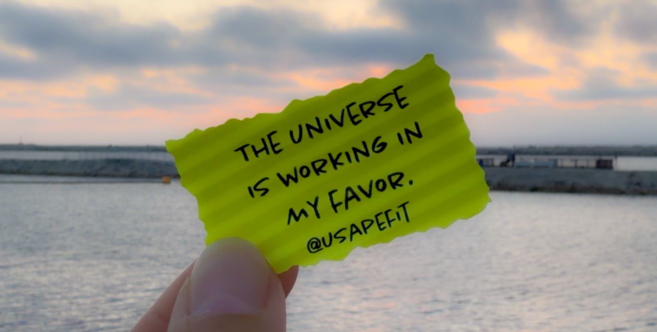 Hand holding a yellow note card with motivational quote 'The Universe is working in my favor' by @usapetit at sunset.