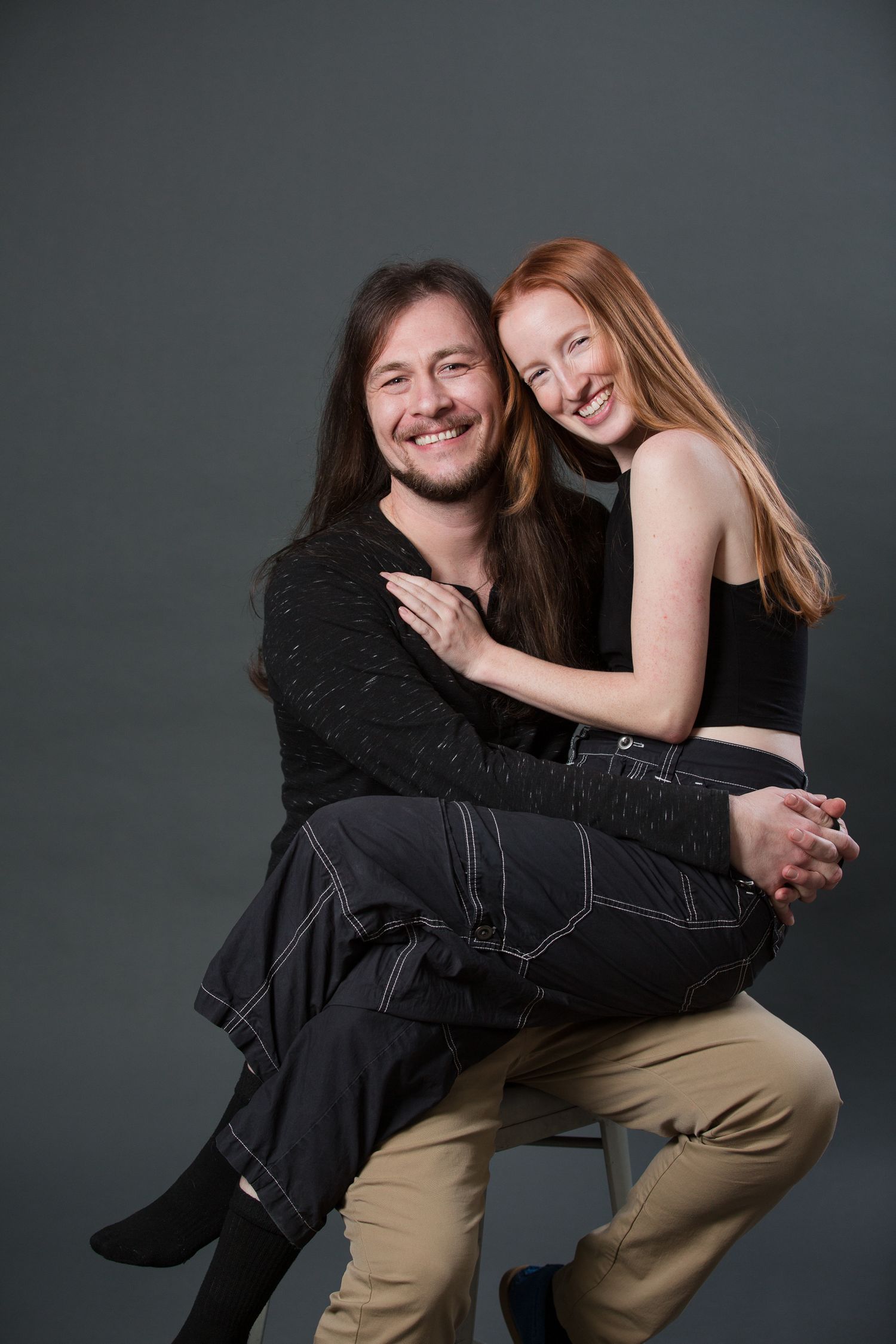 A smiling couple poses together, woman with red hair sitting on man's lap, both wearing black tops in a studio.
