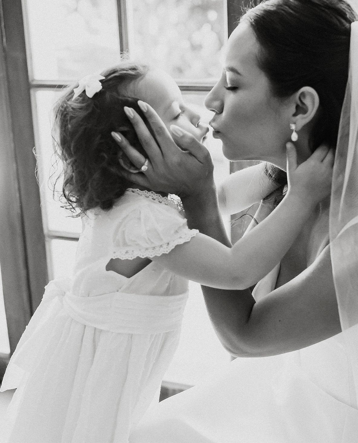 Black and white photo of bride kissing a young flower girl by a sunlit window.