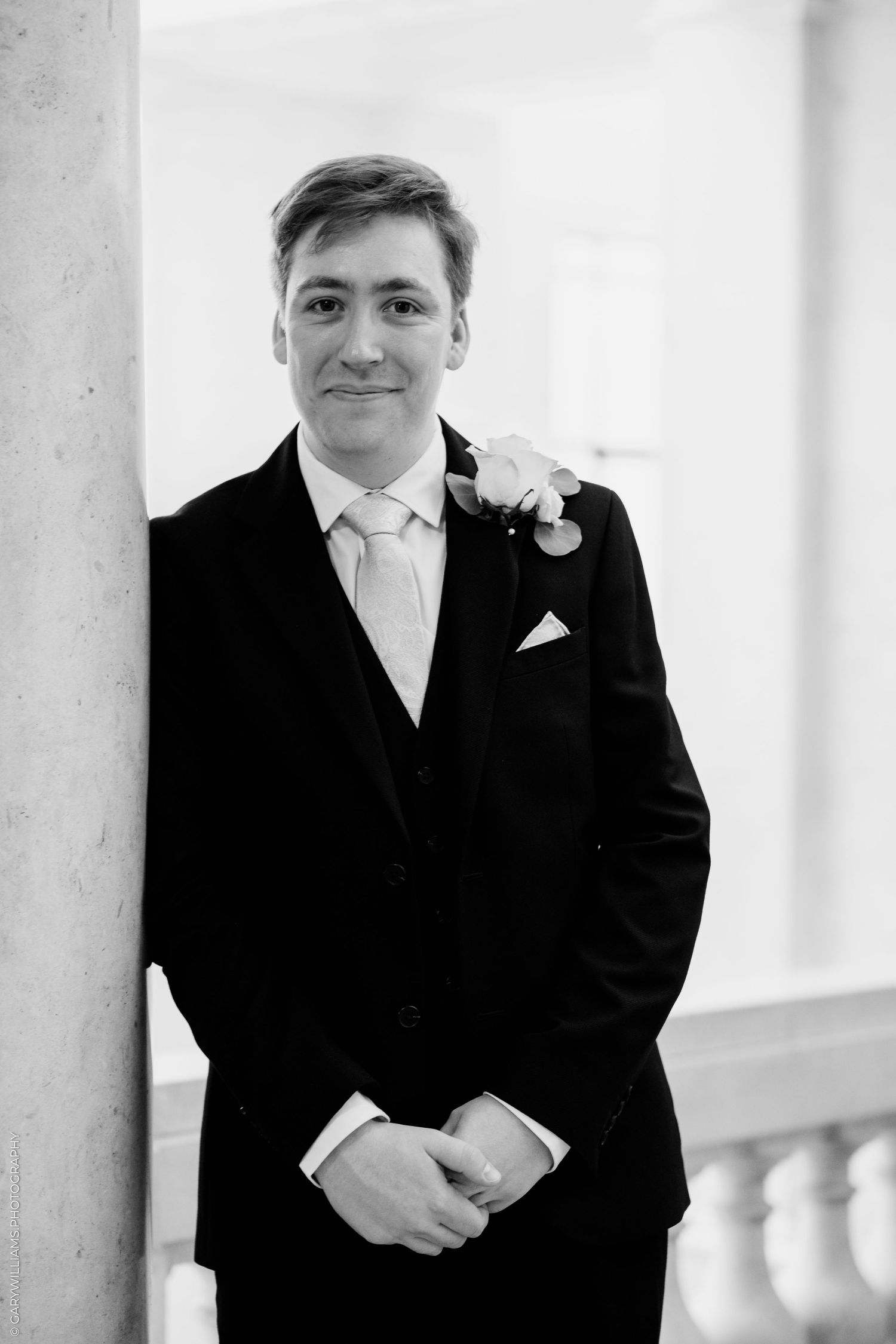 Black and white portrait of a groom in a suit with boutonniere, smiling beside a column.