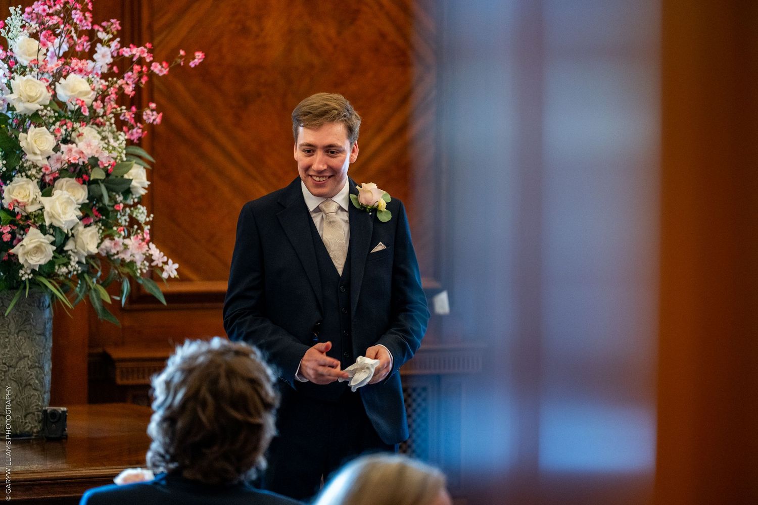 Groom smiling at wedding ceremony altar, wearing navy suit with boutonniere, floral arrangement in background.