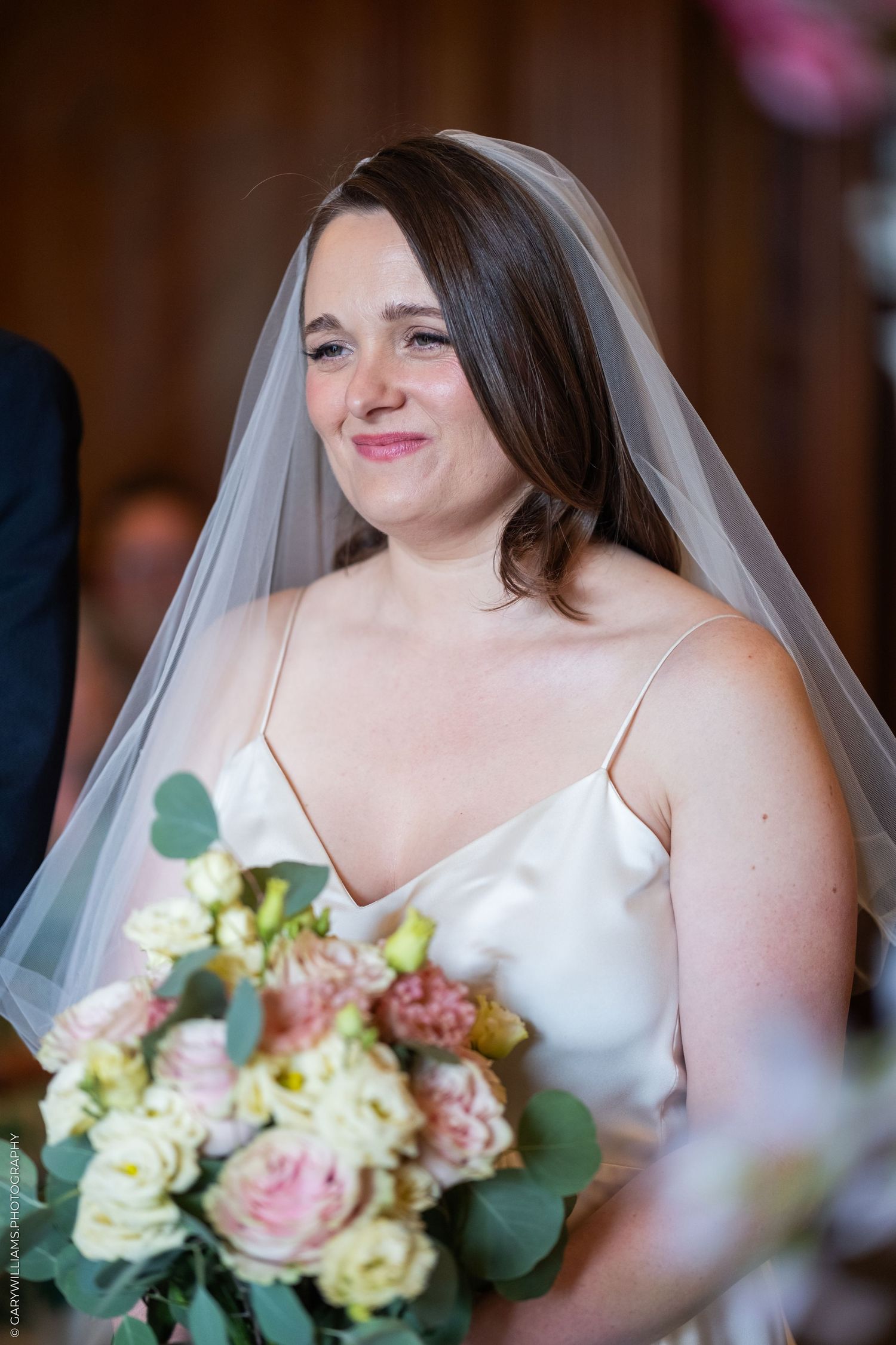 Smiling bride in white satin gown and veil holding pink and cream floral bouquet during wedding ceremony.