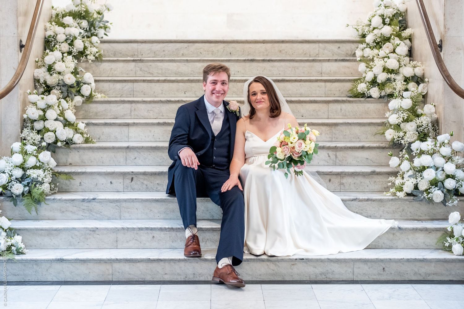 Bride and groom sitting on marble stairs adorned with white floral arrangements, smiling on their wedding day.