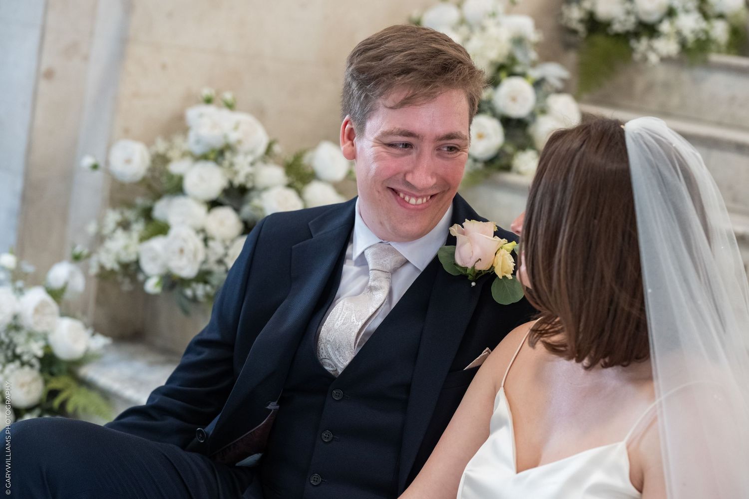 Smiling groom in navy suit looks lovingly at bride in white veil during wedding ceremony.