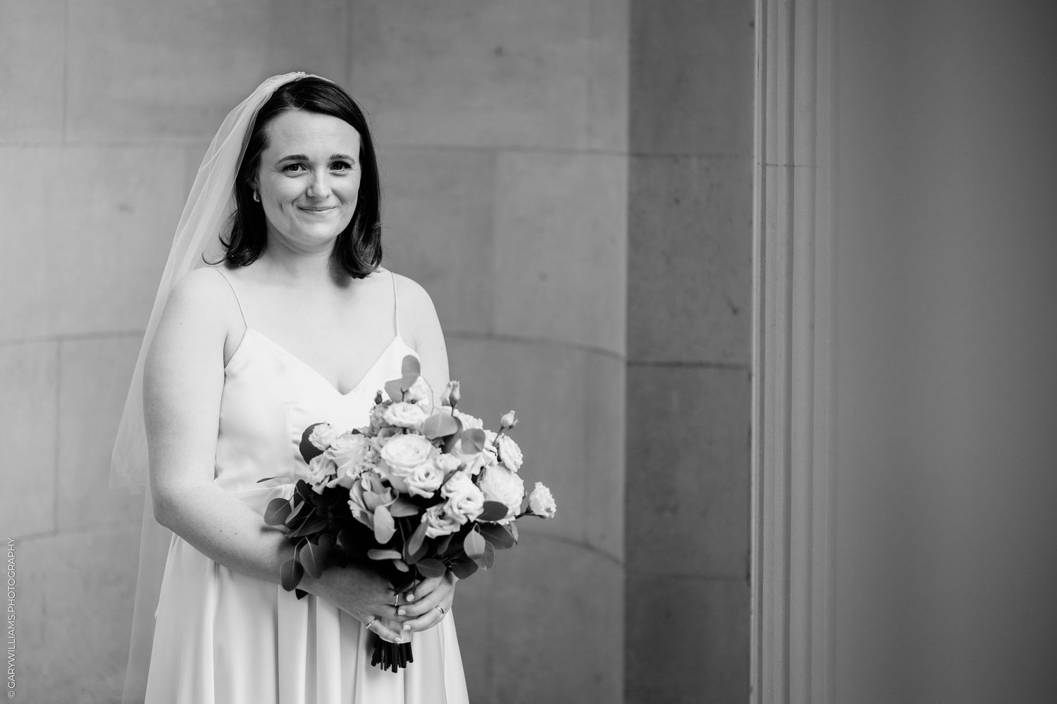 Black and white photo of a smiling bride in a white gown and veil holding a floral bouquet.
