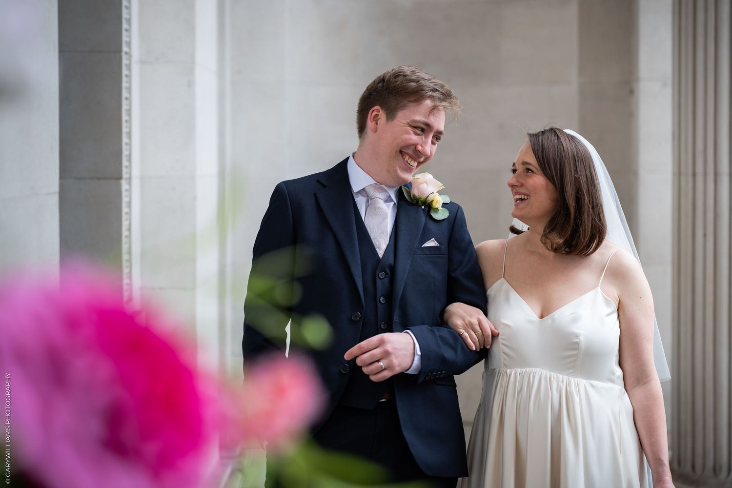 Smiling bride and groom sharing a joyful moment beside elegant stone columns on their wedding day.