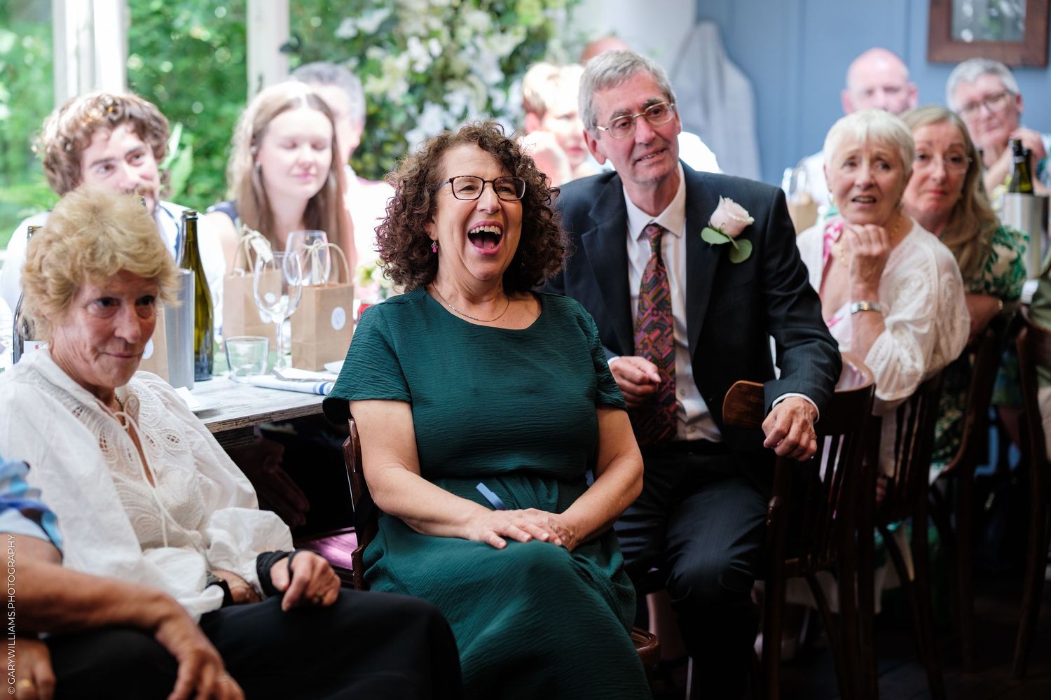 Wedding guests laughing and smiling during a reception speech, seated in a decorated venue.