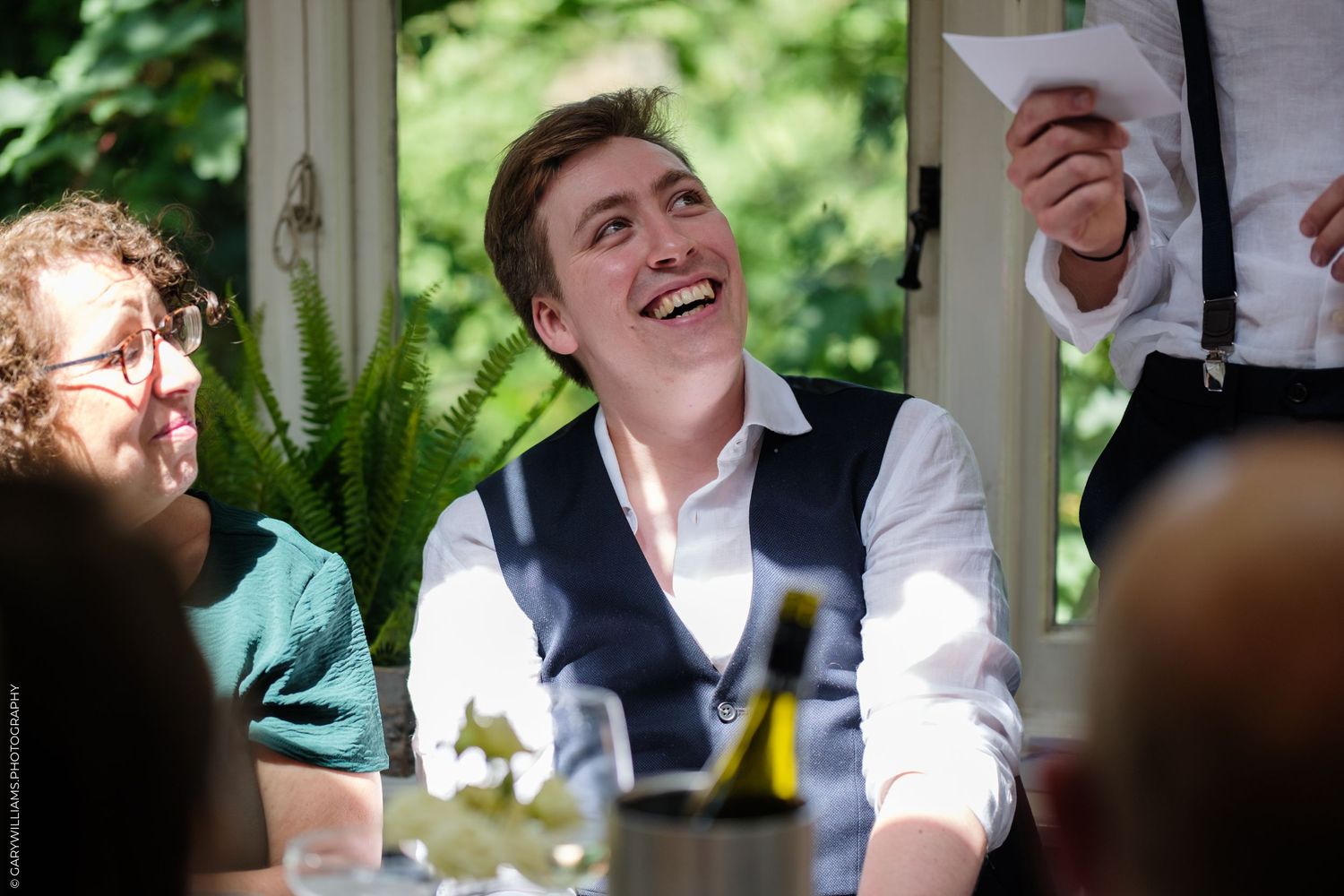 Young man in white shirt and waistcoat laughing joyfully at a wedding reception dinner.