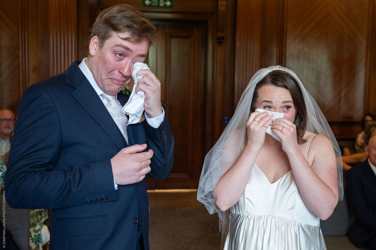 Emotional bride and groom wiping tears with handkerchiefs during their wedding ceremony indoors.