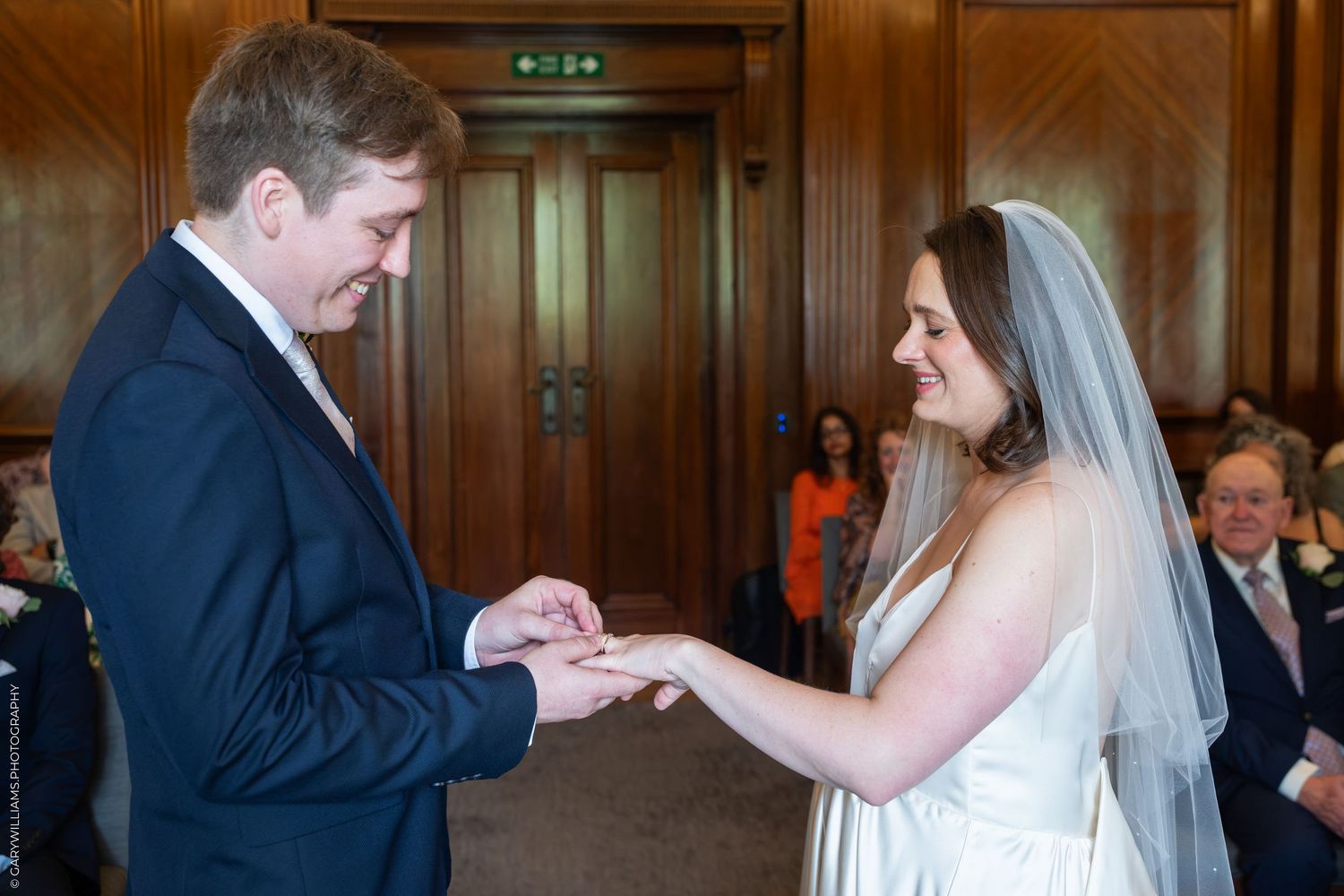 Groom placing wedding ring on brides finger during ceremony at elegant wood-paneled venue.