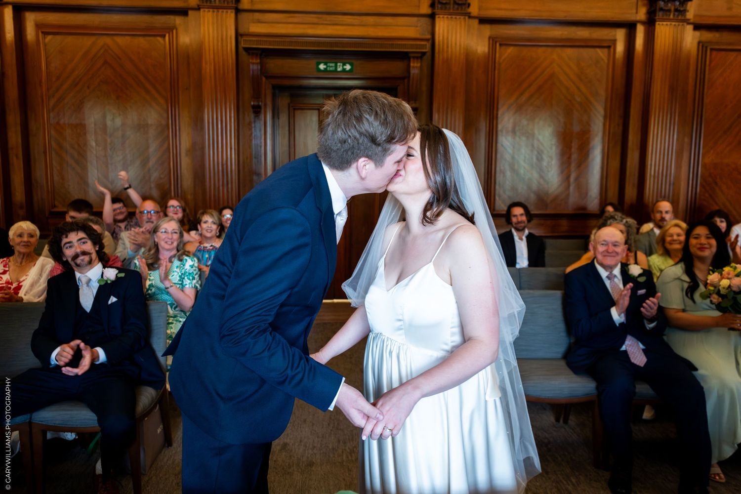 Bride and groom share first kiss at wedding ceremony while guests cheer and applaud in elegant venue.