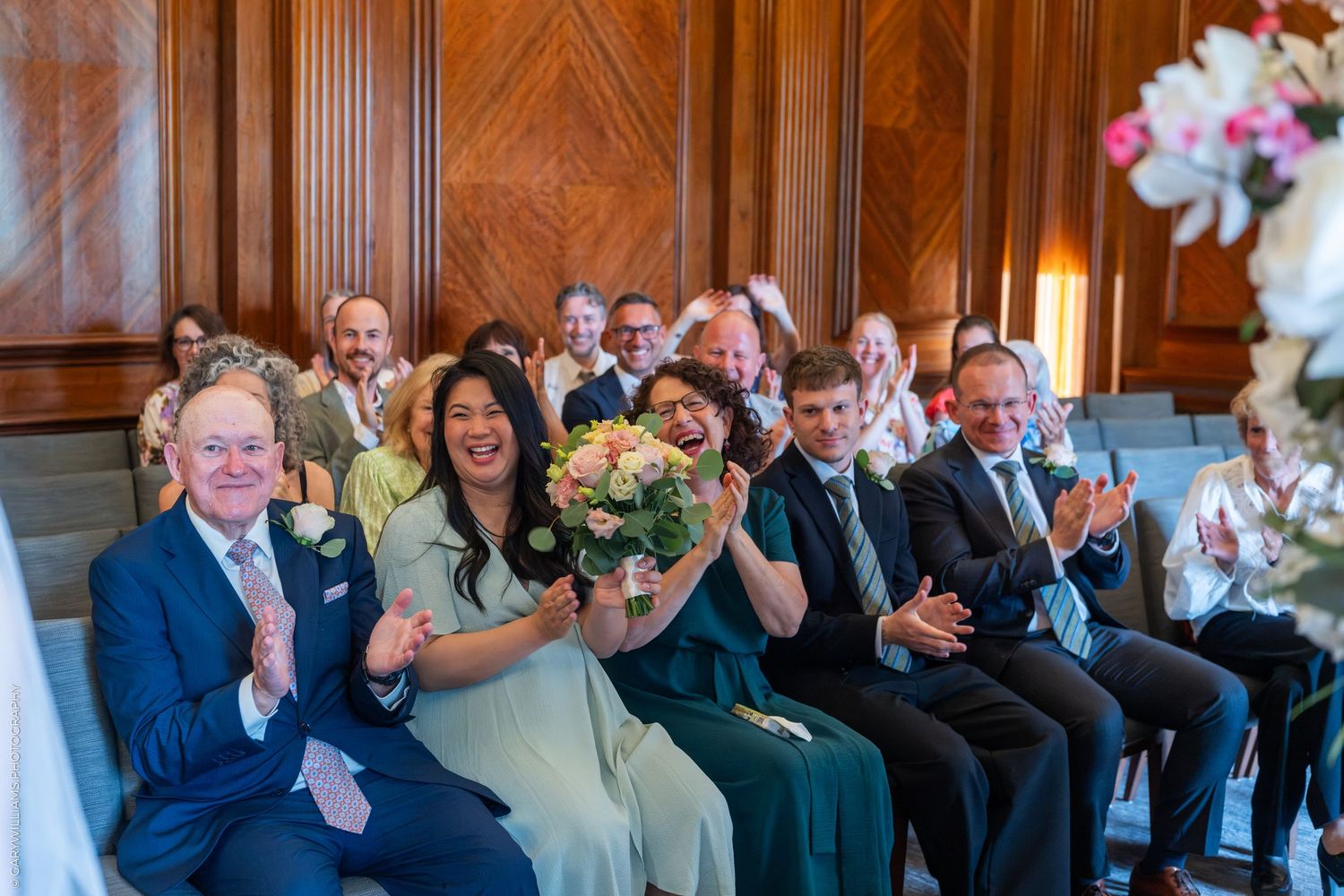 Joyful wedding guests clapping and smiling during a ceremony in an elegant wood-paneled room, bride holding a pastel bouquet.