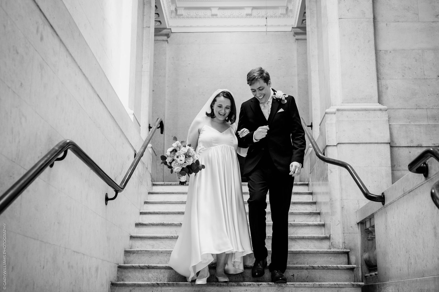 Black and white photo of a bride and groom walking down grand staircase, bride holding bouquet, both smiling.