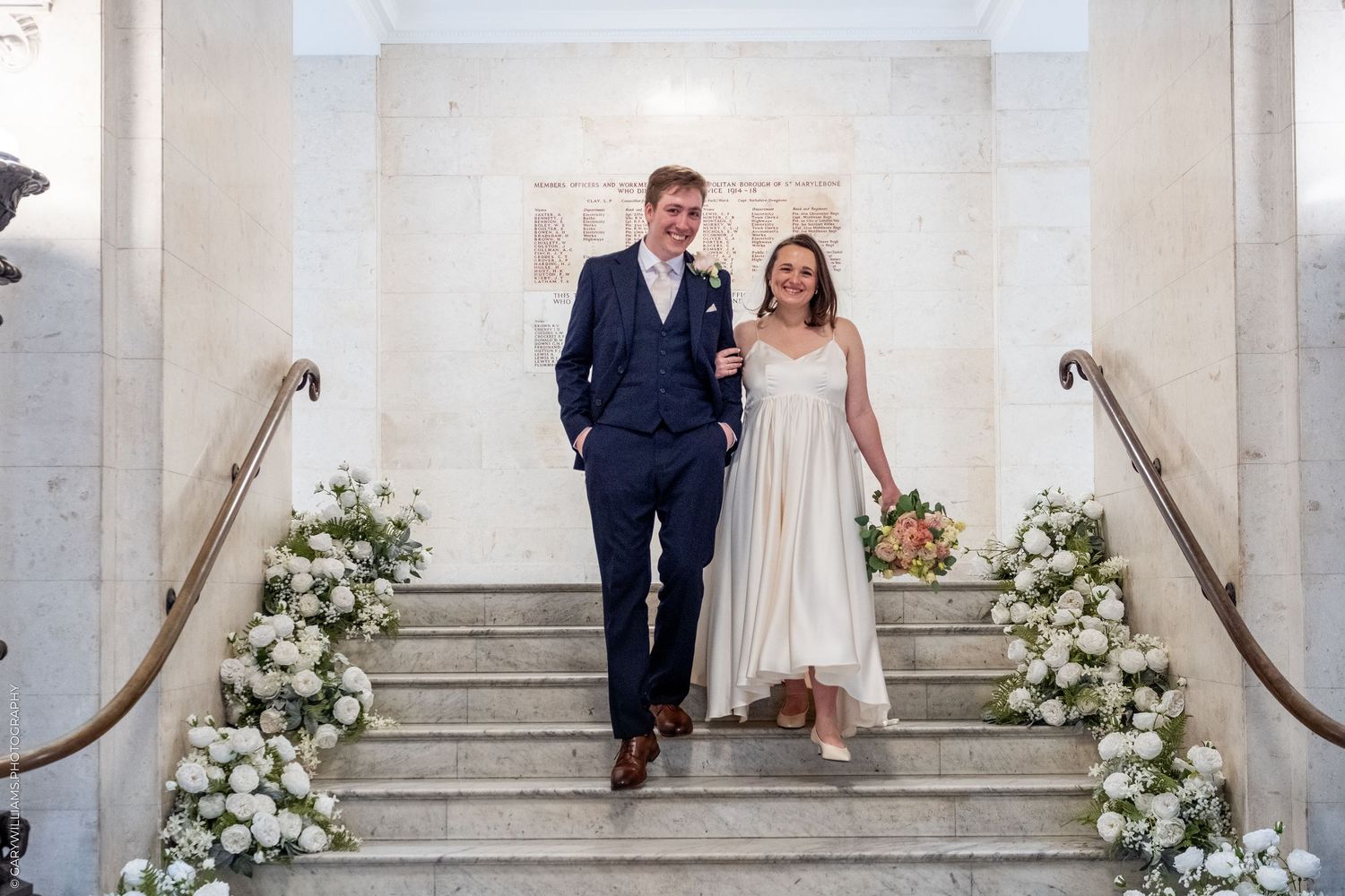 Bride and groom descend floral-decorated staircase, bride in tea-length dress, groom in navy suit, at elegant wedding venue.