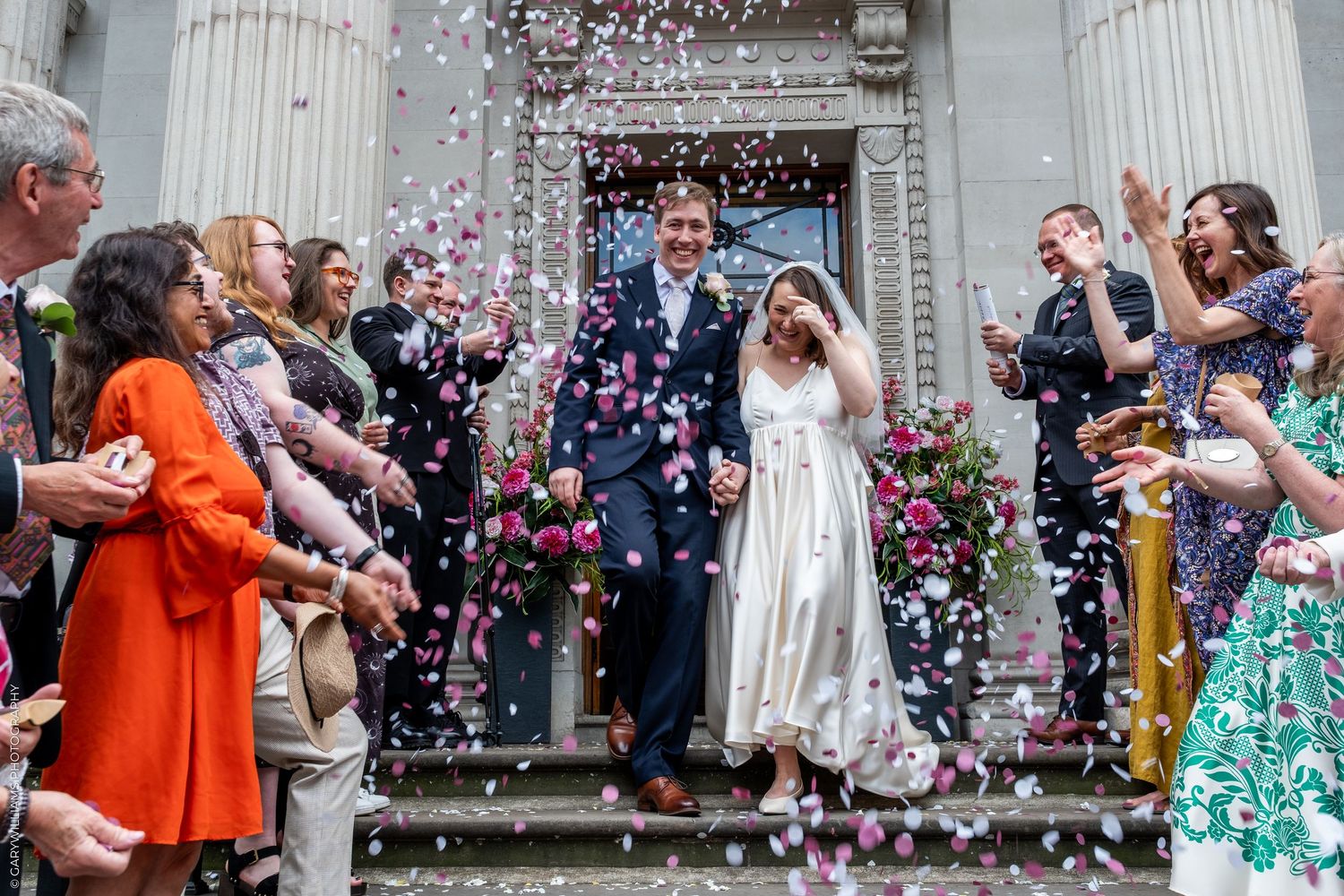 Bride and groom walk through colorful confetti shower as wedding guests celebrate outside a grand building.