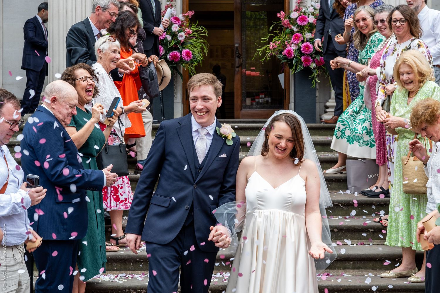 Newlywed couple walks through confetti shower on church steps surrounded by cheering wedding guests on a sunny day.