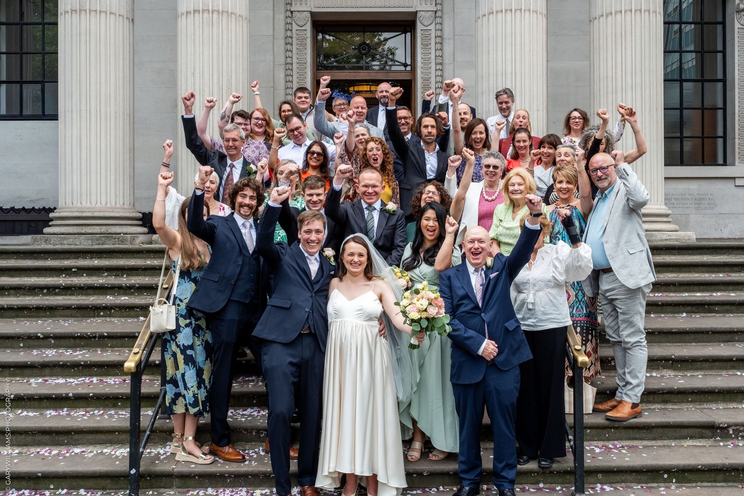 Large wedding group celebrating on steps, guests cheering with raised fists, bride holding bouquet in white dress.
