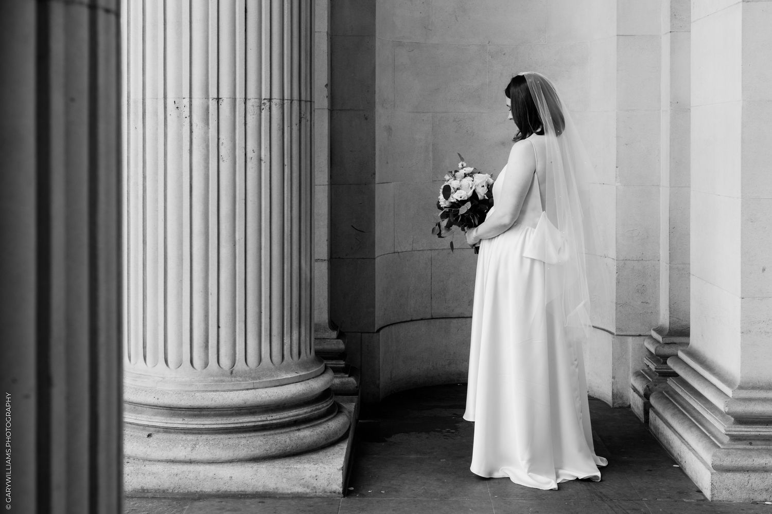 Black and white photo of a bride in a white gown holding a bouquet, standing between classical stone columns.