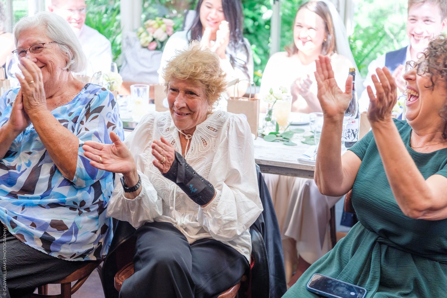 Three elderly women clapping and laughing joyfully at a celebratory event indoors.