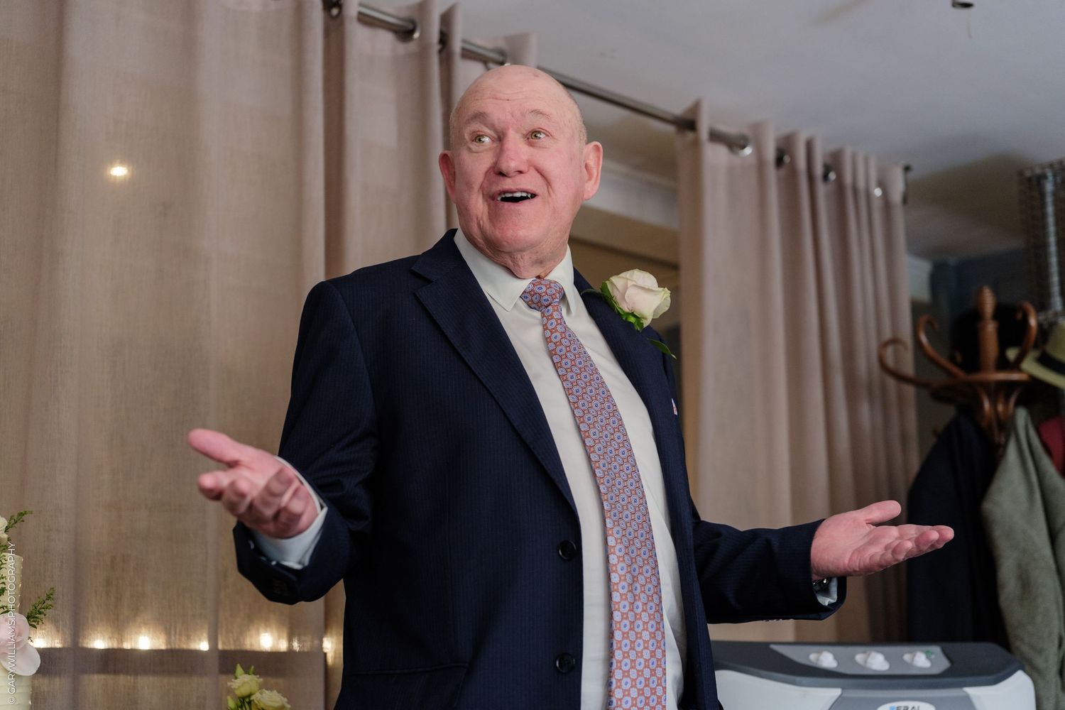 Elderly man in a suit gesturing with open hands while speaking at an indoor event.