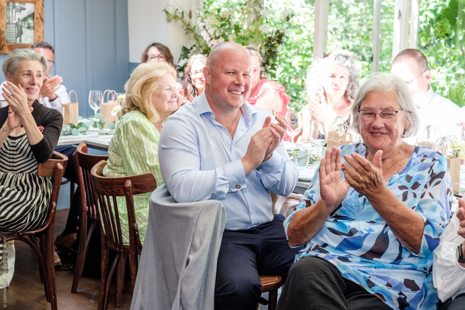 Group of smiling people clapping at an indoor celebration, including elderly women and a tall man in a blue shirt.