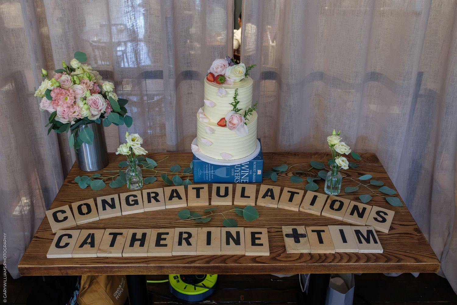 Wedding cake display with Scrabble tiles spelling Congratulations Catherine and Tim on a rustic wooden table with flowers.
