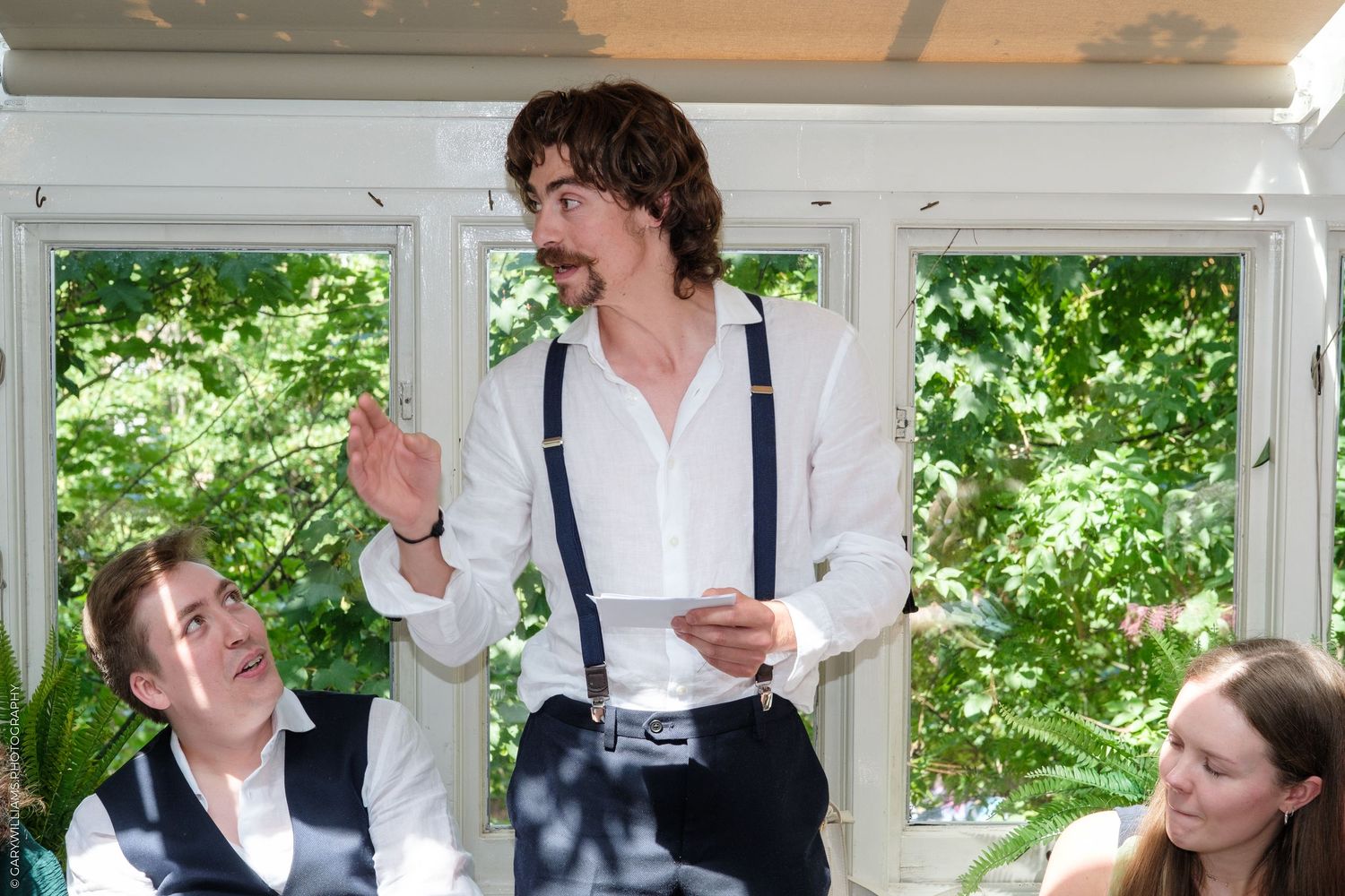 Man in white shirt and suspenders giving a speech at a wedding reception, gesturing while two guests listen attentively.