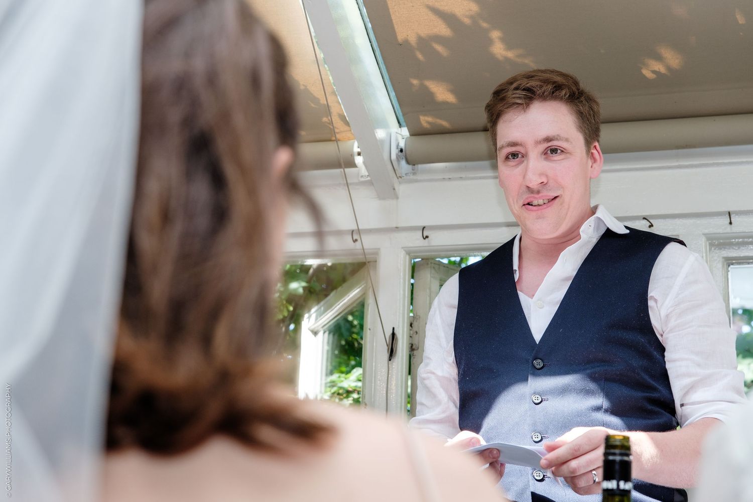 Man in navy vest speaking to bride in wedding veil at outdoor ceremony, holding a card.