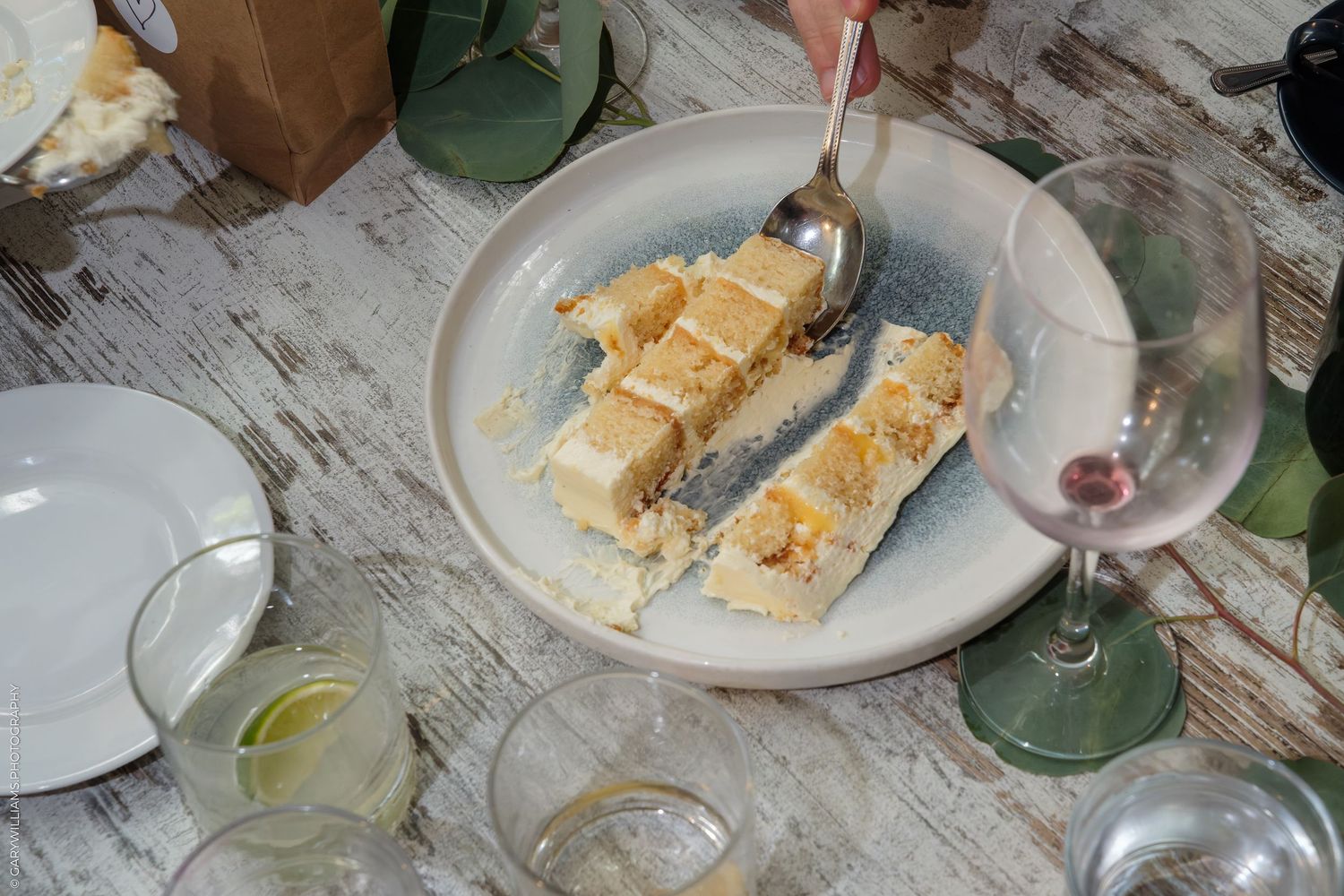A hand holding a spoon over two slices of layered cake on a ceramic plate at a rustic dining table.