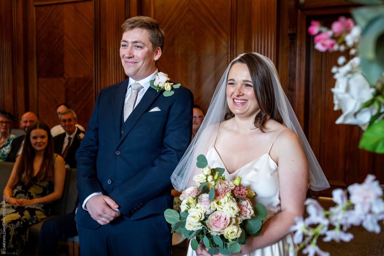 Bride holding pink floral bouquet and groom in navy suit smiling during wedding ceremony indoors.