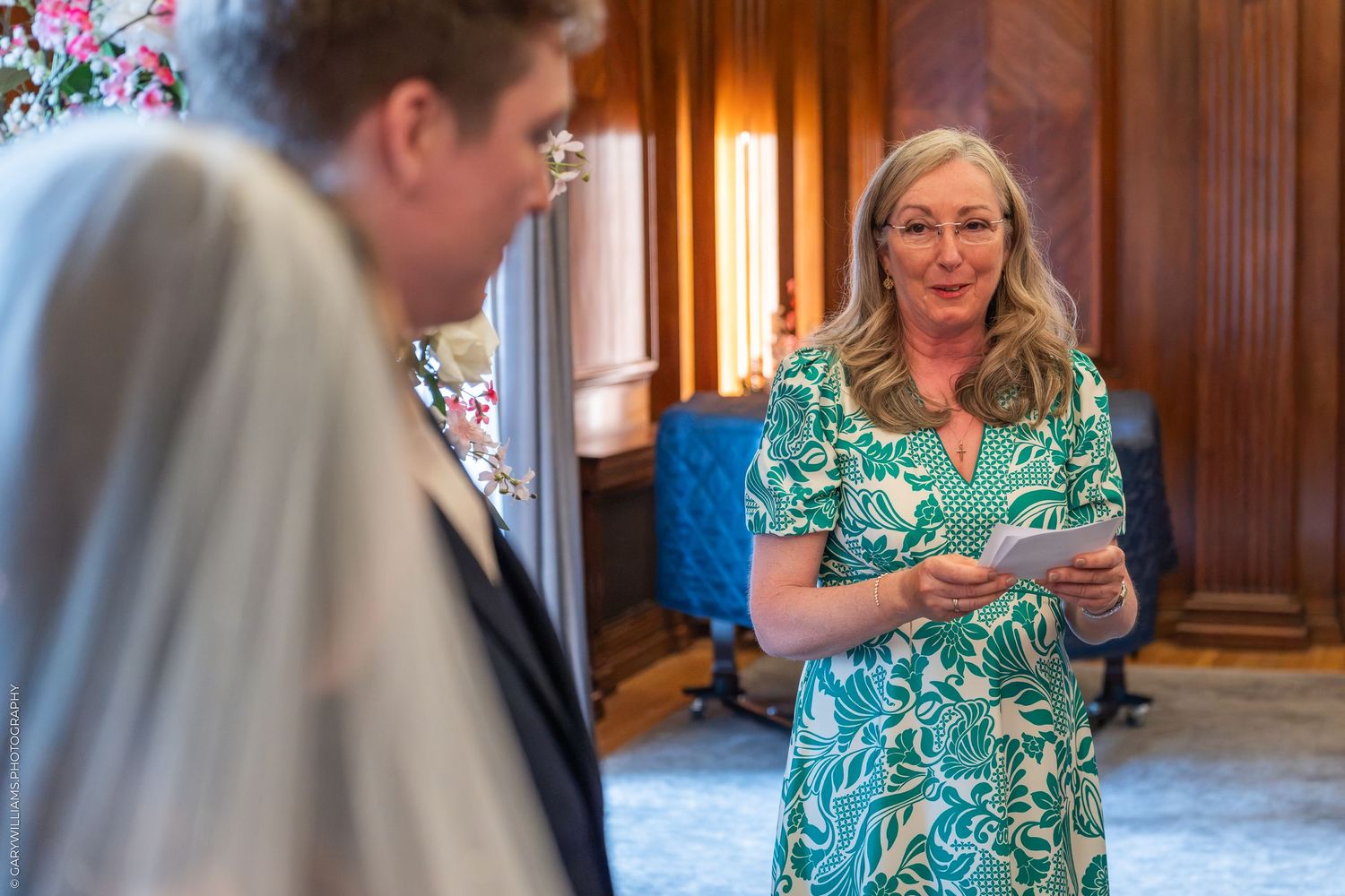 Woman in green floral dress reading vows during an elegant indoor wedding ceremony.