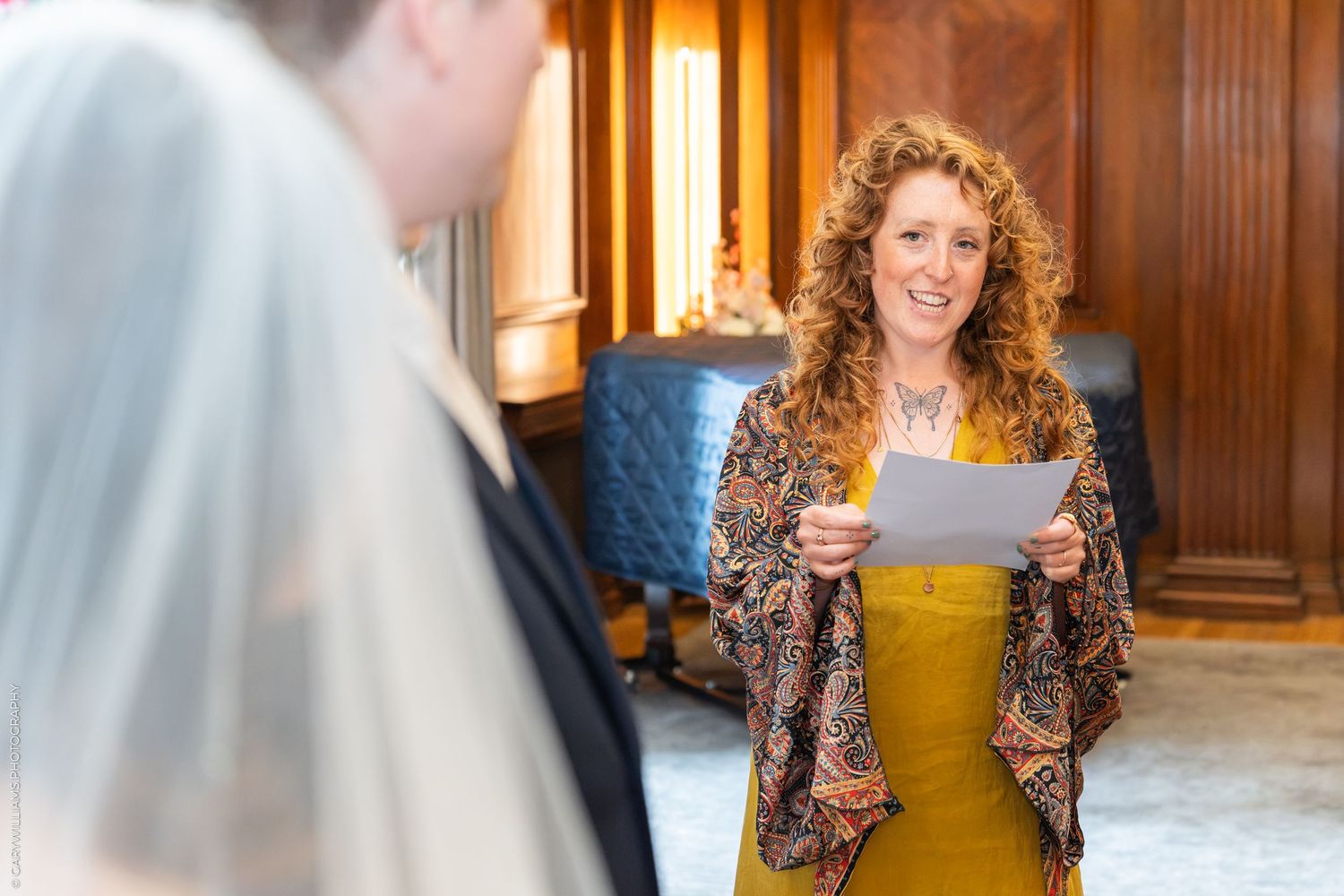 Smiling wedding officiant with curly red hair reads from paper during an indoor ceremony in an elegant room.