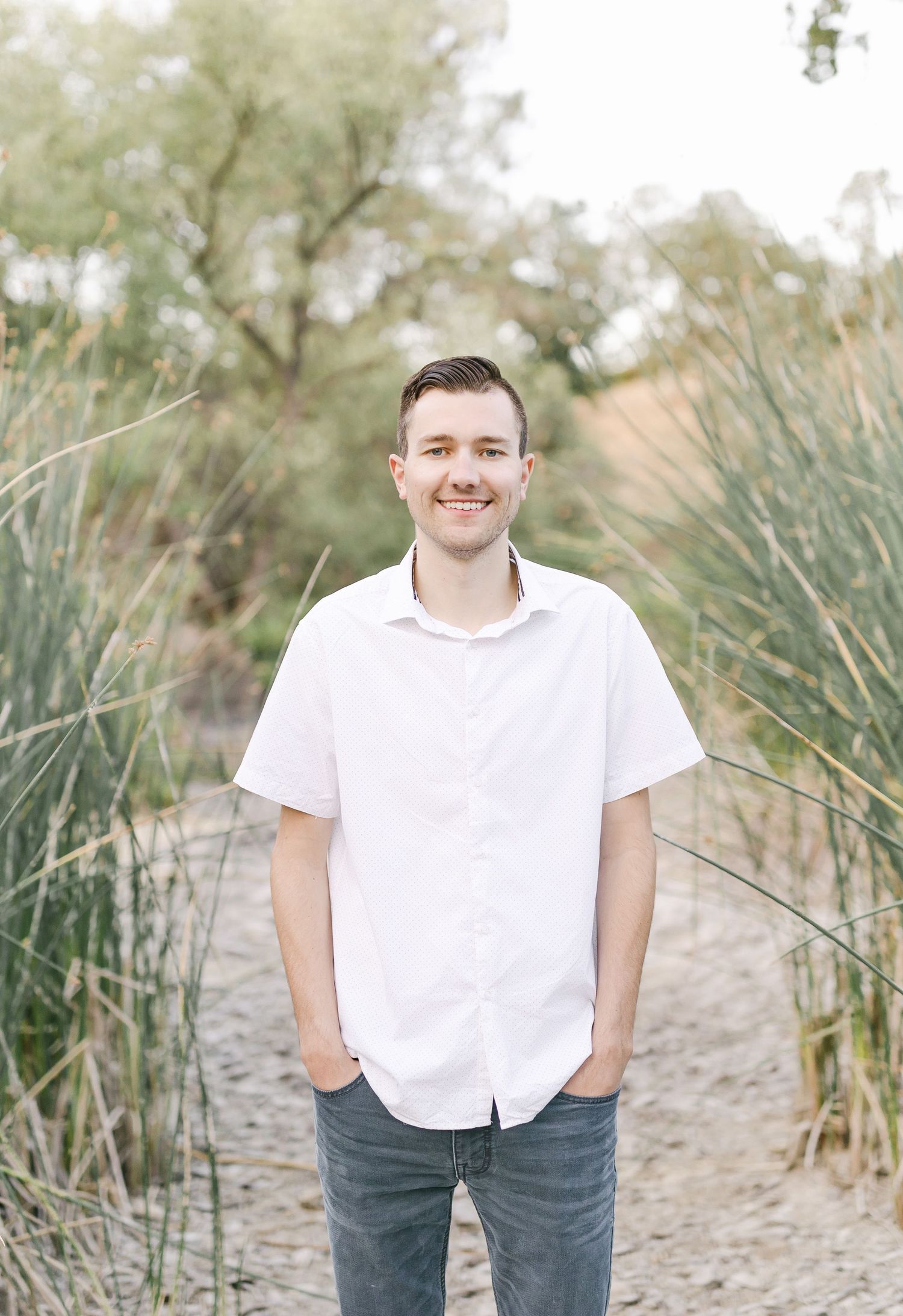 A smiling young man in a white button-up shirt and jeans stands on a sandy path surrounded by tall grass and trees.