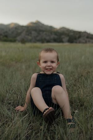 Smiling toddler boy sitting in green grass field wearing denim overalls with rocky mountain hills in background.