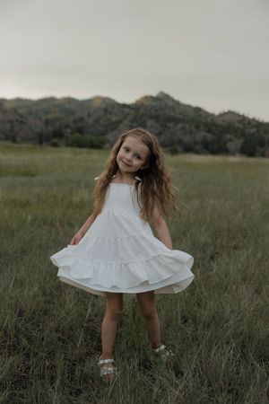 A young girl in a white tiered dress twirls in a green meadow with mountains in the background.