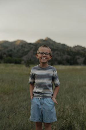 Smiling boy in glasses and striped shirt stands in grassy mountain field.