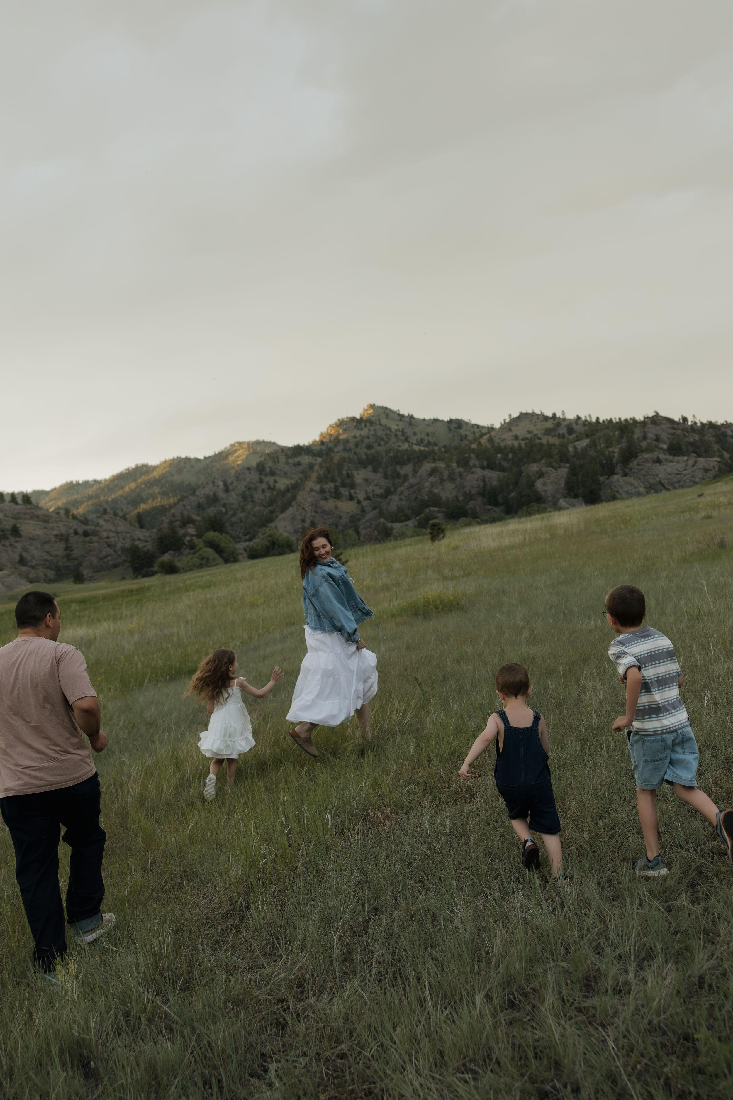 Family running through grassy hillside field at dusk with mountain backdrop, mother turning to smile.