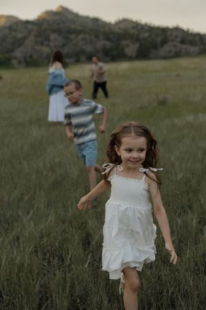 Young girl in white dress runs through green meadow with family and rocky hills in background.