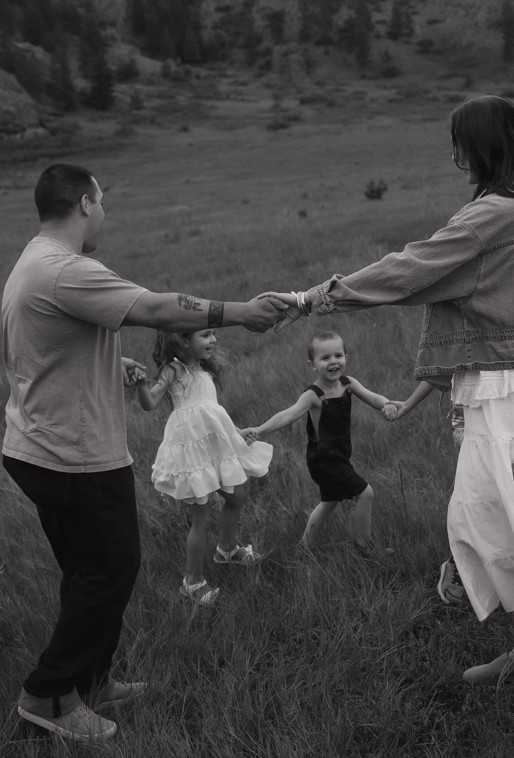 Black and white photo of a joyful family holding hands in a circle, swinging two young girls in a grassy field.