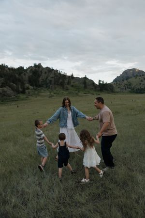 Family of five holding hands in circle on green meadow with rocky mountain landscape at twilight.