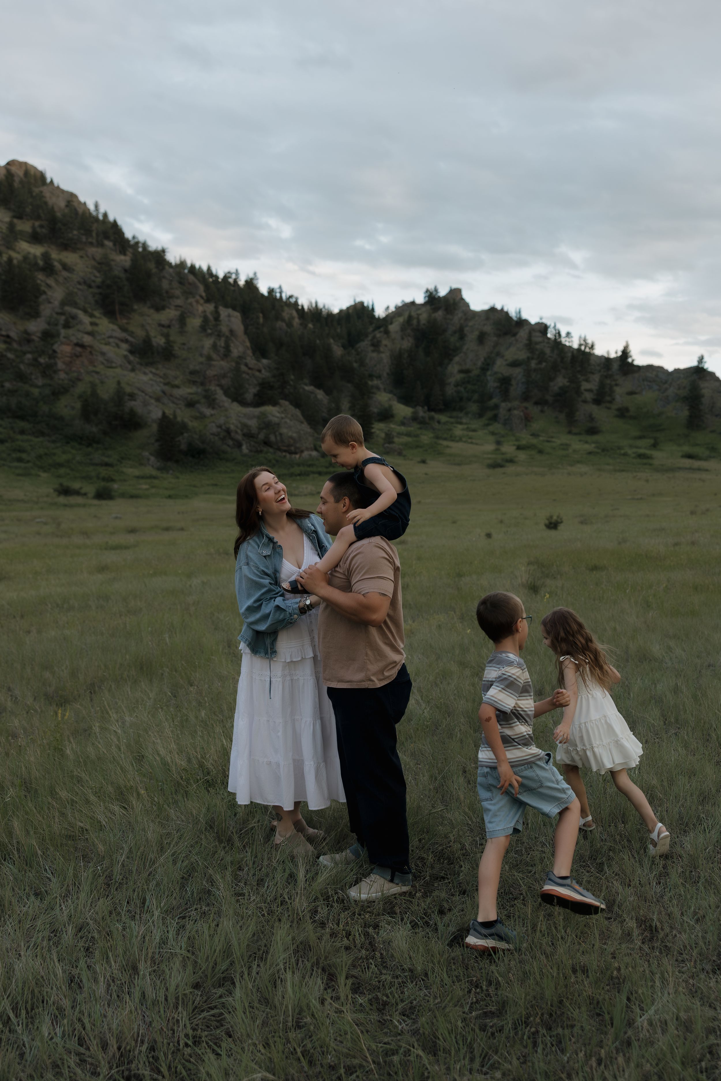 Family of five laughing together in a green mountain meadow at dusk, parents embracing while children run toward them.
