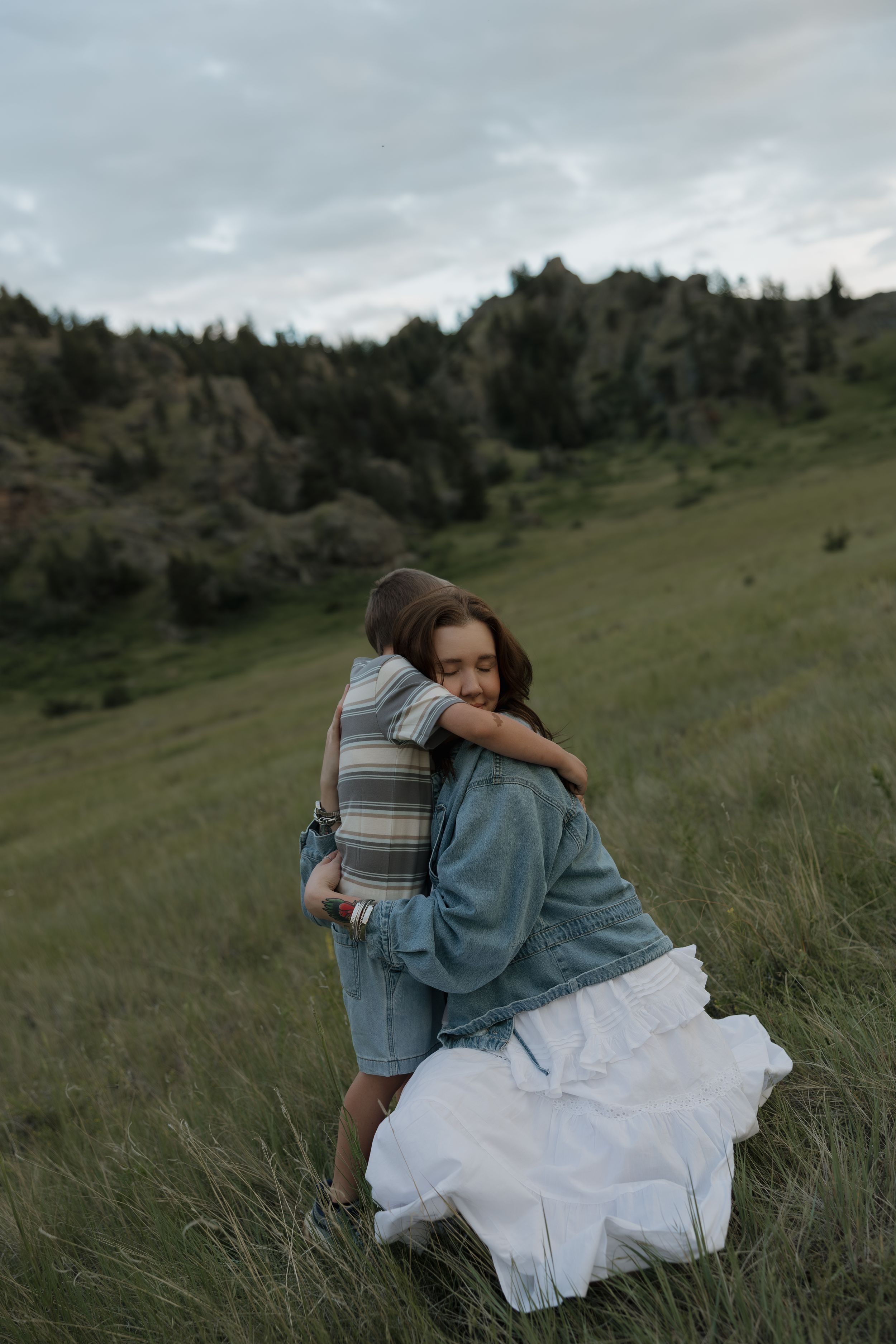 Mother in denim jacket and white skirt hugs child in grassy field with rocky hills behind.