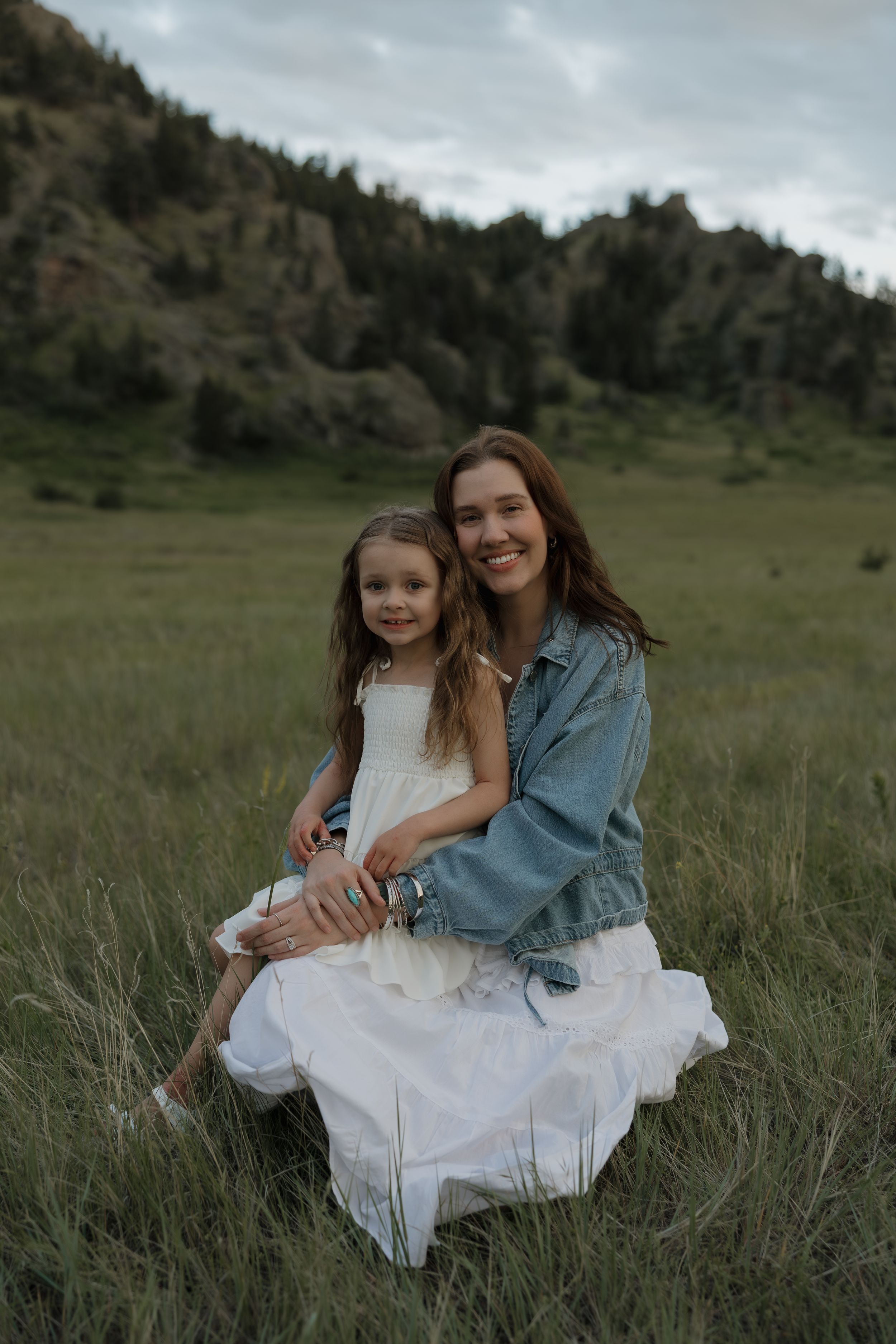 Smiling mother in denim jacket and white dress holds young daughter in grassy mountain field at dusk.