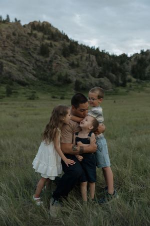 A father kneels in a mountain meadow, embraced by three young children in a heartwarming outdoor family portrait.