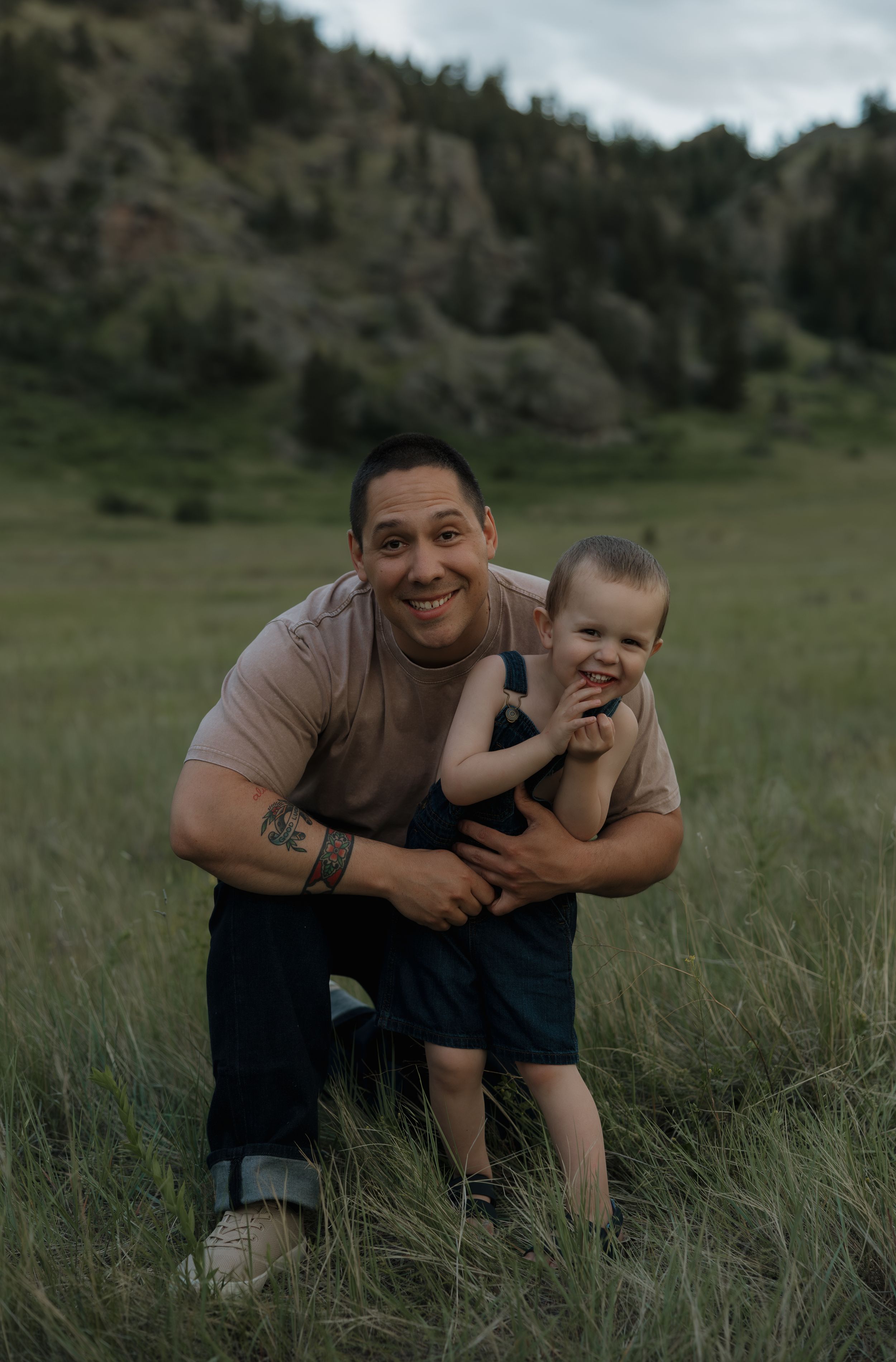 A smiling man kneels in a grassy mountain meadow hugging a happy toddler boy during an outdoor family photo session.