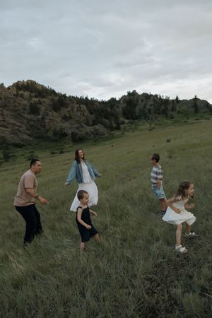Family of five running through a green meadow with rocky hills and trees in the background on a cloudy day.