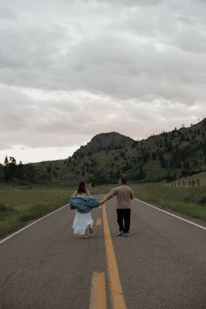 Couple holding hands walking down an empty mountain road at dusk in a scenic green valley.