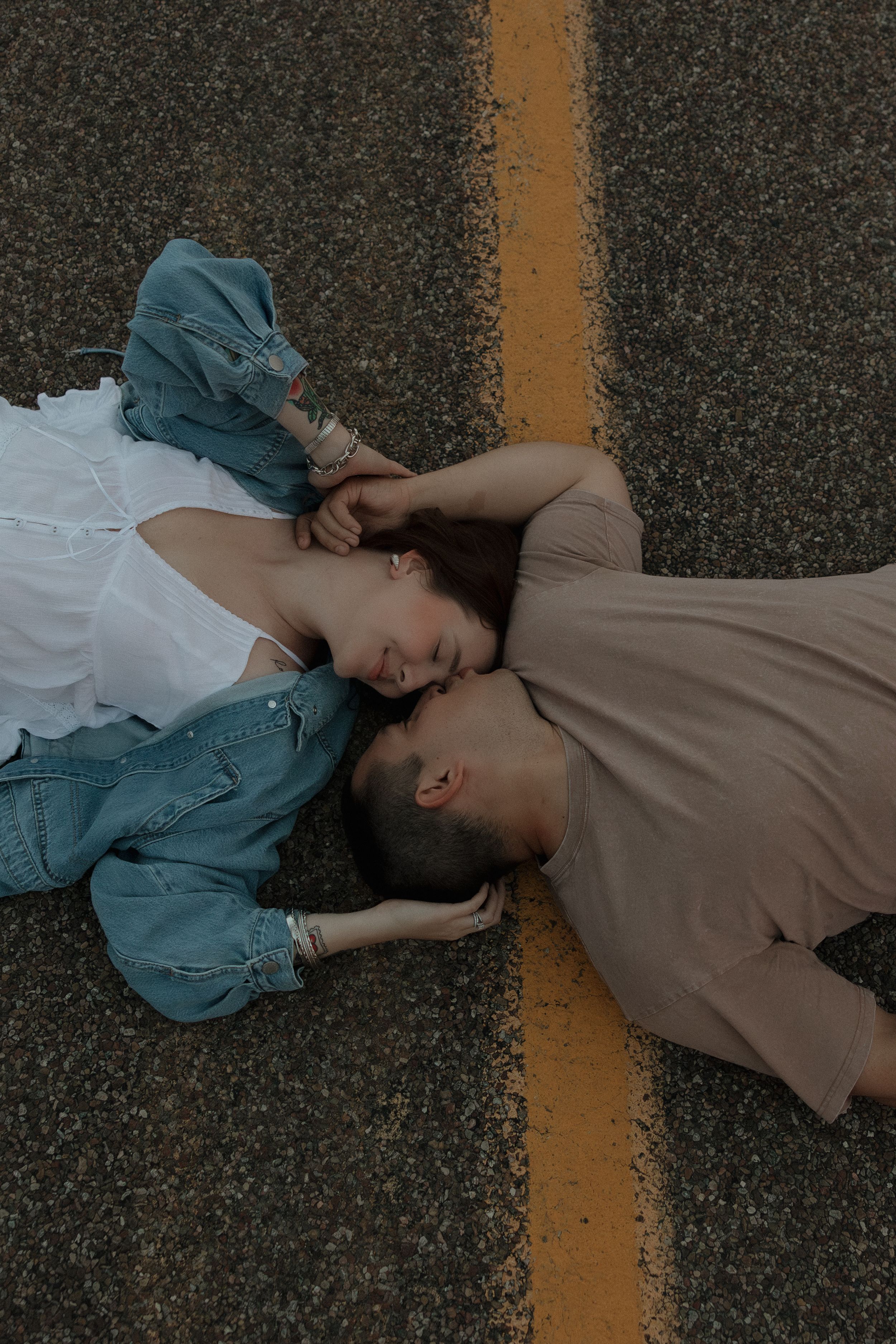 A romantic couple lies on a road beside a yellow line, facing each other tenderly during an outdoor engagement photo shoot.