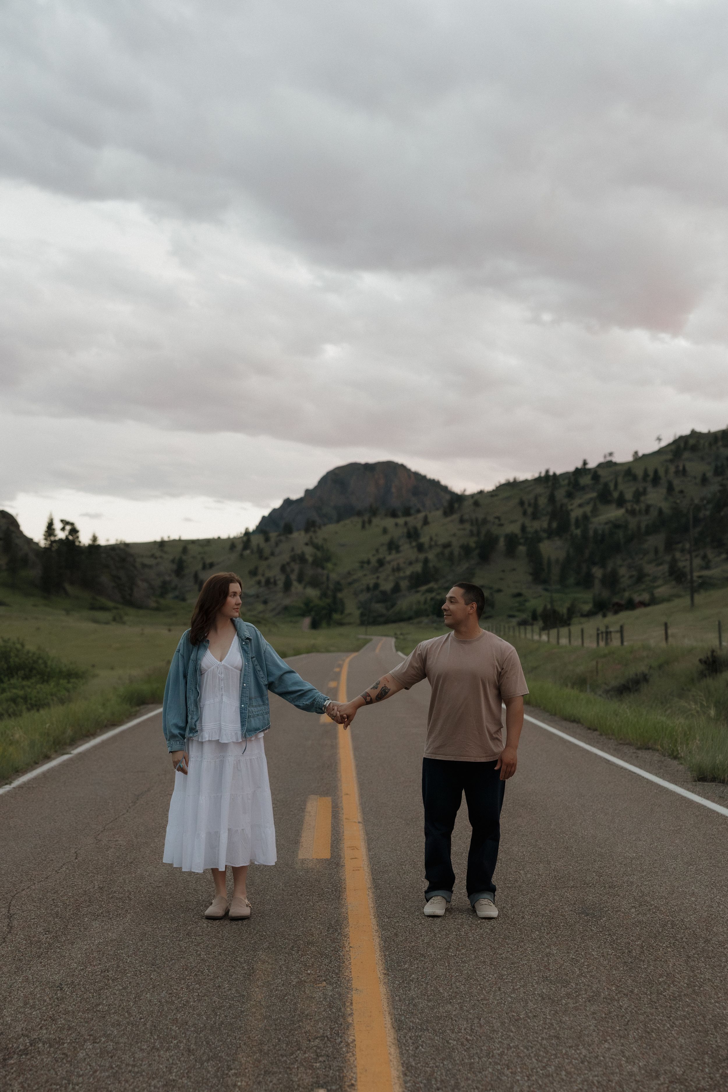 Couple holds hands on empty road with green hills and cloudy sky.