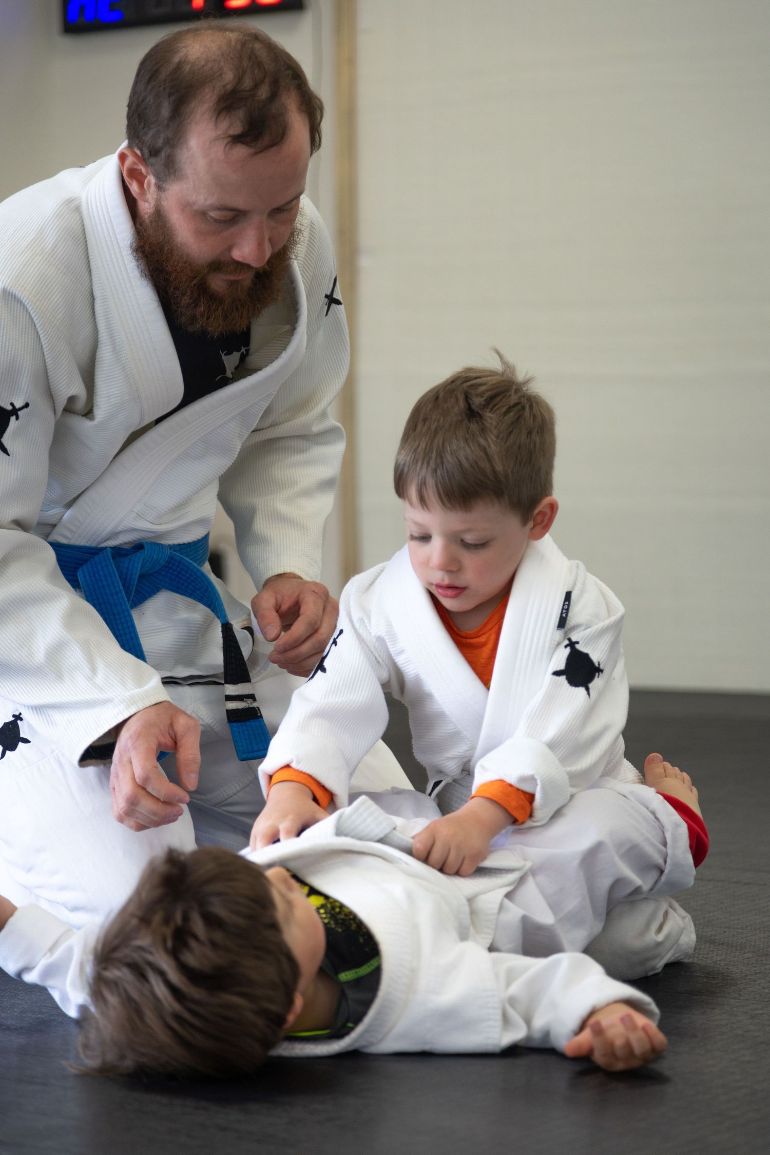 Adult BJJ instructor in white gi guides two young children practicing Brazilian jiu-jitsu techniques on a mat.